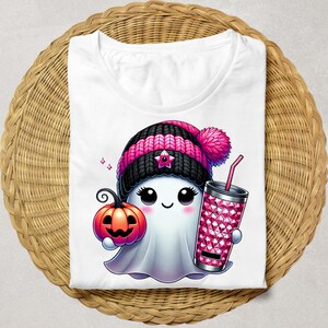 May include: A white t-shirt with a cartoon ghost wearing a pink and black knit hat with a pom-pom. The ghost is holding a pumpkin and a pink glittery tumbler with a straw.
