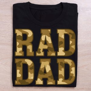 May include: Black t-shirt with the words "RAD DAD" in large, block letters. The letters are filled with a camouflage pattern in shades of brown and tan. The shirt is folded neatly on a light wood surface.