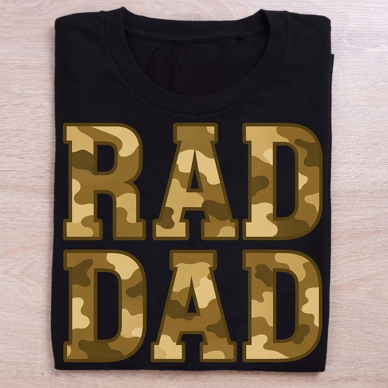 May include: Black t-shirt with the words "RAD DAD" in large, block letters. The letters are filled with a camouflage pattern in shades of brown and tan. The shirt is folded neatly on a light wood surface.
