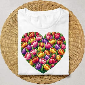 Pode incluir: Camiseta branca com um design em forma de cora&ccedil;&atilde;o composto por tulipas coloridas. As tulipas s&atilde;o em tons de vermelho, rosa, amarelo e roxo, com folhas verdes. A camiseta est&aacute; dobrada sobre uma superf&iacute;cie tecida marrom claro.
