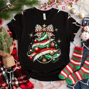 May include: Black t-shirt with a Christmas tree design. The tree is decorated with a leopard print bow, red and green ornaments, and white glitter.