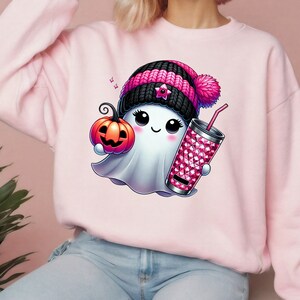 May include: A pink sweatshirt with a cute cartoon ghost wearing a pink and black knit hat, holding a pumpkin and a pink sparkly cup with a straw.