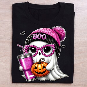 K&ouml;nnte beinhalten: Ein schwarzes T-Shirt mit einem rosa und schwarzen Geist, der eine rosa M&uuml;tze mit dem Wort "BOO" tr&auml;gt. Der Geist h&auml;lt einen K&uuml;rbis und einen rosa Glitzerbecher mit einem Strohhalm.
