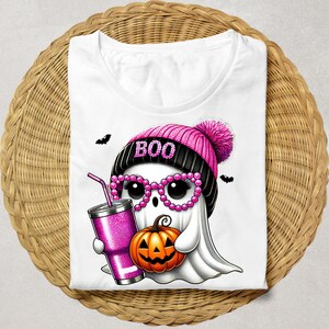 K&ouml;nnte beinhalten: Wei&szlig;es T-Shirt mit einem rosa und schwarzen Geist, der eine rosa M&uuml;tze mit dem Wort "BOO" tr&auml;gt. Der Geist h&auml;lt einen K&uuml;rbis und einen rosa Glitzerbecher mit einem Strohhalm.