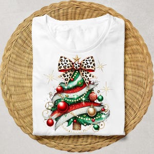 May include: White t-shirt with a Christmas tree design. The tree is green with red and white stripes, gold glitter, and a leopard print bow. The tree is decorated with gold and red ornaments.