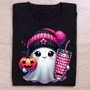 May include: A black t-shirt with a white ghost wearing a pink and black knit hat with a star on it. The ghost is holding a pink pumpkin and a pink and white glittery cup with a straw.