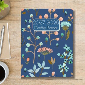May include: A rectangular monthly planner with a dark blue cover featuring a botanical pattern of light blue, pink, and teal flowers and berries. The text 2027-2028 Monthly Planner is printed in white within a rectangular frame on the front cover.