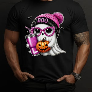 K&ouml;nnte beinhalten: Ein schwarzes T-Shirt mit einem rosa und wei&szlig;en Geist, der eine rosa M&uuml;tze mit dem Wort "BOO" tr&auml;gt. Der Geist h&auml;lt einen K&uuml;rbis und einen rosa Glitzerbecher mit einem Strohhalm.