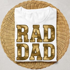 May include: White t-shirt with the words "RAD DAD" in large, block letters. The letters have a camouflage pattern in shades of brown and tan. The shirt is displayed on a round, woven wicker surface.