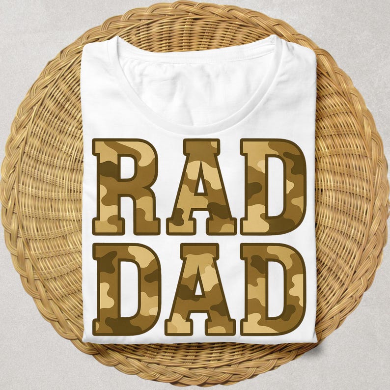 May include: White t-shirt with the words "RAD DAD" in large, block letters. The letters have a camouflage pattern in shades of brown and tan. The shirt is displayed on a round, woven wicker surface.