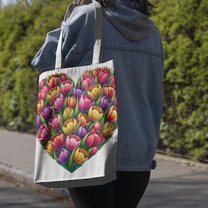 Pode incluir: Uma sacola branca com um design em forma de cora&ccedil;&atilde;o preenchido com tulipas coloridas. As tulipas s&atilde;o em tons de vermelho, rosa, amarelo e roxo, com folhas verdes. A sacola &eacute; carregada no ombro.