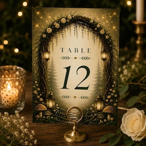May include: A table number card with a whimsical forest theme. The card features the word "TABLE" above the number "12" in a dark green font. The design includes a floral arch, lanterns, and mushroom illustrations. The card is held by a gold-colored stand.