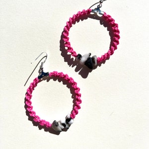 以下が含まれることがあります： 黒と白の石のアクセントが付いたピンクのフープイヤリングのペア。イヤリングは編まれたコードで作られており、銀色のフックが付いています。
