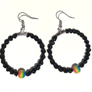 May include: A pair of black macrame hoop earrings with silver-colored hooks. Each earring features a rainbow-colored bead. The earrings are displayed against a white background.