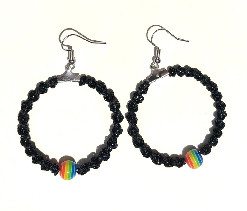 May include: A pair of black macrame hoop earrings with silver-colored hooks. Each earring features a rainbow-colored bead. The earrings are displayed against a white background.