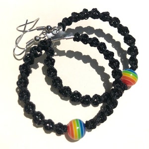 May include: Black macrame hoop earrings with a rainbow-striped bead accent. The earrings have silver-colored ear wires. The macrame is knotted in a spiral pattern. The earrings are displayed on a white surface.