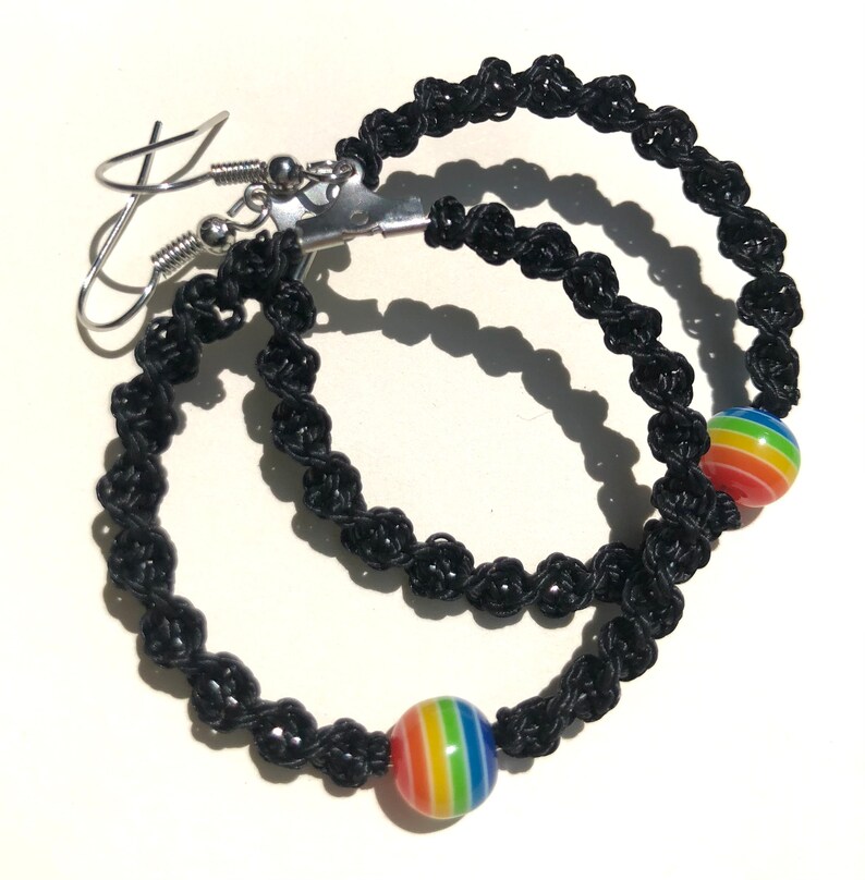 May include: Black macrame hoop earrings with a rainbow-striped bead accent. The earrings have silver-colored ear wires. The macrame is knotted in a spiral pattern. The earrings are displayed on a white surface.