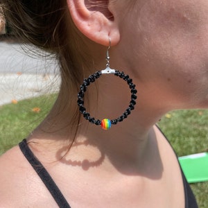 May include: Black macrame hoop earrings with a silver-tone hook. A small, round bead with rainbow colors is attached to the bottom of the hoop. The earrings are worn on a person.