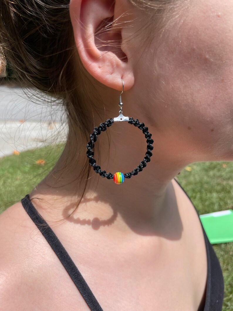 May include: Black macrame hoop earrings with a silver-tone hook. A small, round bead with rainbow colors is attached to the bottom of the hoop. The earrings are worn on a person.