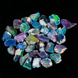 May include: A collection of rough-cut opal gemstones, showcasing a vibrant display of colors including blues, greens, pinks, and purples. The opals are set against a black background, highlighting their iridescent qualities. These natural stones vary in size and shape.