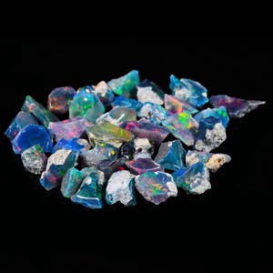 May include: A collection of raw opal gemstones, displaying a variety of colors including blue, green, pink, and iridescent flashes. The rough stones have a natural, unpolished appearance, with a dark background.