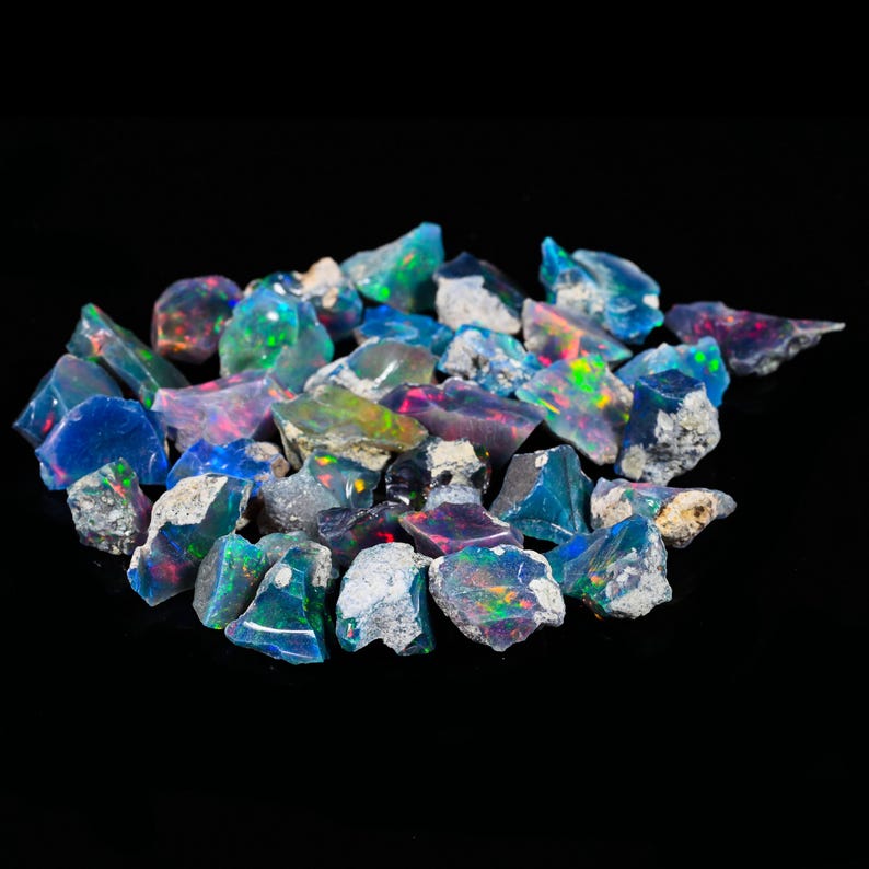 May include: A collection of raw opal gemstones, displaying a variety of colors including blue, green, pink, and iridescent flashes. The rough stones have a natural, unpolished appearance, with a dark background.