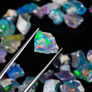 May include: A close-up of a vibrant opal gemstone held by silver tweezers, showcasing a play of color with green, blue, and red hues. Other rough opal stones are scattered in the background against a black surface.