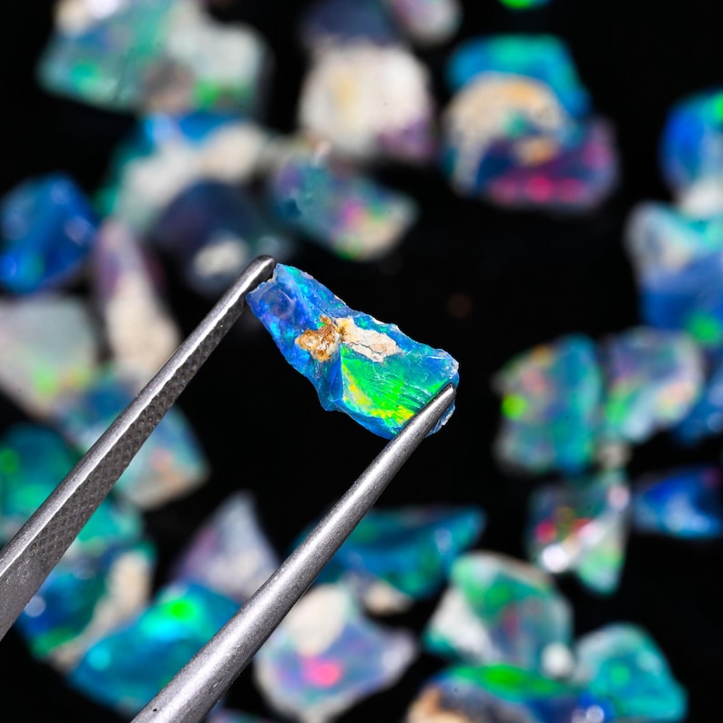 May include: A close-up of a vibrant opal gemstone held by tweezers, showcasing a spectrum of colors including blue, green, and yellow. The rough-cut stone has a unique, natural shape. Other opals are blurred in the background.