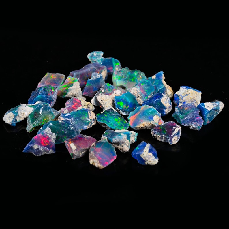 May include: A collection of raw opal gemstones. The opals display a variety of colors, including blue, green, red, and purple, with a shimmering, iridescent effect. The stones have a rough, natural texture and are set against a black background.