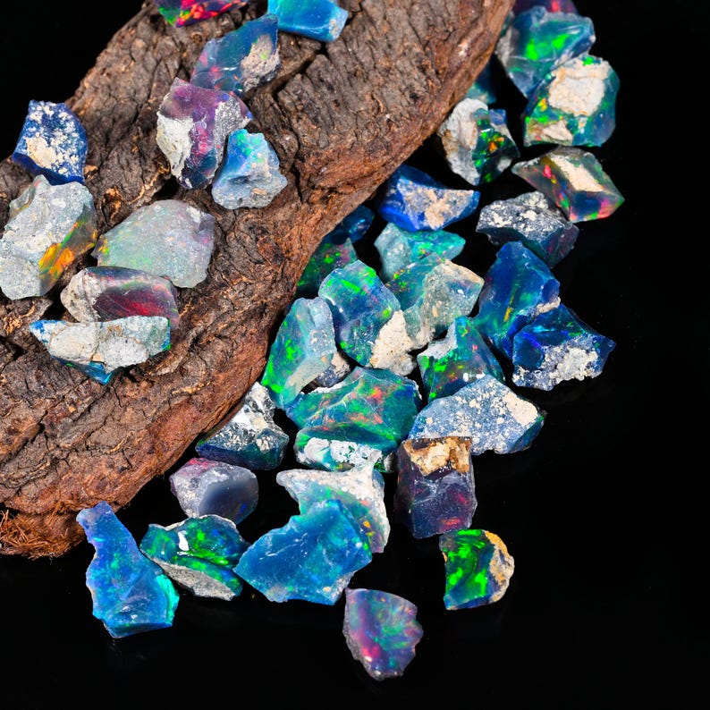 May include: A collection of raw opal gemstones in various shades of blue, green, and purple, with flashes of color. The opals are scattered on and around a piece of dark brown wood, set against a black background.