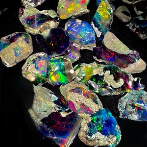 Puede incluir: Una colección de piedras preciosas de ópalo en bruto, que muestra una vibrante gama de colores que incluyen azules, verdes, rojos y amarillos. Los ópalos tienen una textura rugosa y natural y se colocan sobre un fondo negro, resaltando sus cualidades iridiscentes.