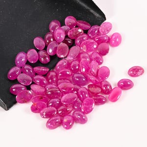 Op de afbeelding: Een verzameling ovaalvormige, gepolijste robijn edelstenen in verschillende tinten roze en rood. De edelstenen liggen verspreid op een witte ondergrond, met een zwarte schep in de linkerbovenhoek. De edelstenen hebben een gladde, glanzende afwerking.