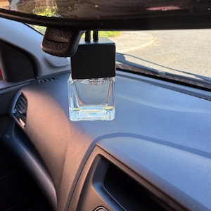 May include: A clear, rectangular car air freshener with a black top hangs from a car's rearview mirror. The air freshener is suspended above a dark dashboard, with a glimpse of the outside world visible through the windshield.