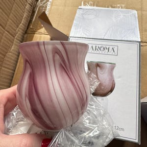 May include: A decorative glass vase with a swirling pattern of white and mauve. The vase is in a box with the text "AROMA ACCESSORIES" and a measurement of 12cm. The vase is partially wrapped in plastic.