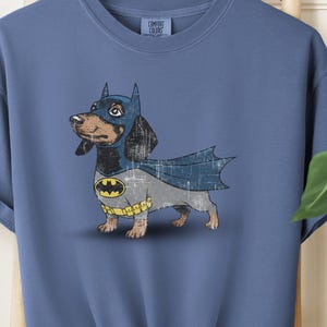 May include: Blue jean-colored t-shirt featuring a cartoon dachshund dressed as Batman. The dog wears a blue cowl, cape, and a yellow and black bat symbol on its chest. The shirt is knotted at the bottom.