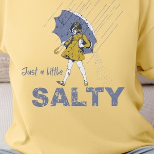 May include: Butter-colored t-shirt with the text "Just a little SALTY" in blue and a graphic of a person under an umbrella. The person is wearing a yellow dress and holding a blue umbrella. The shirt is a casual, unisex style.