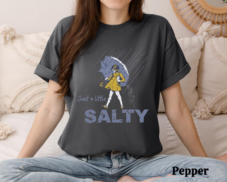 May include: Dark grey t-shirt with a graphic of a person under an umbrella in the rain. The person is wearing a yellow dress and holding a blue umbrella. The text "Just a little SALTY" is below the graphic.