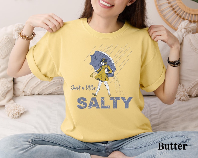 May include: Butter-colored t-shirt with the text "Just a little SALTY" in blue and a graphic of a person under an umbrella. The person is wearing a yellow dress and holding a blue umbrella. The shirt is a casual, unisex style.
