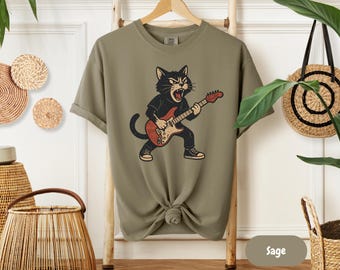 May include: Sage green t-shirt featuring a cartoon cat playing an electric guitar. The cat is black with a red and tan guitar. The shirt has a crew neck and short sleeves. The shirt is knotted at the bottom.