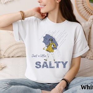 May include: White t-shirt with a graphic of a person under an umbrella in the rain. The text "Just a little SALTY" is below the graphic. The word "White" is printed at the bottom right.