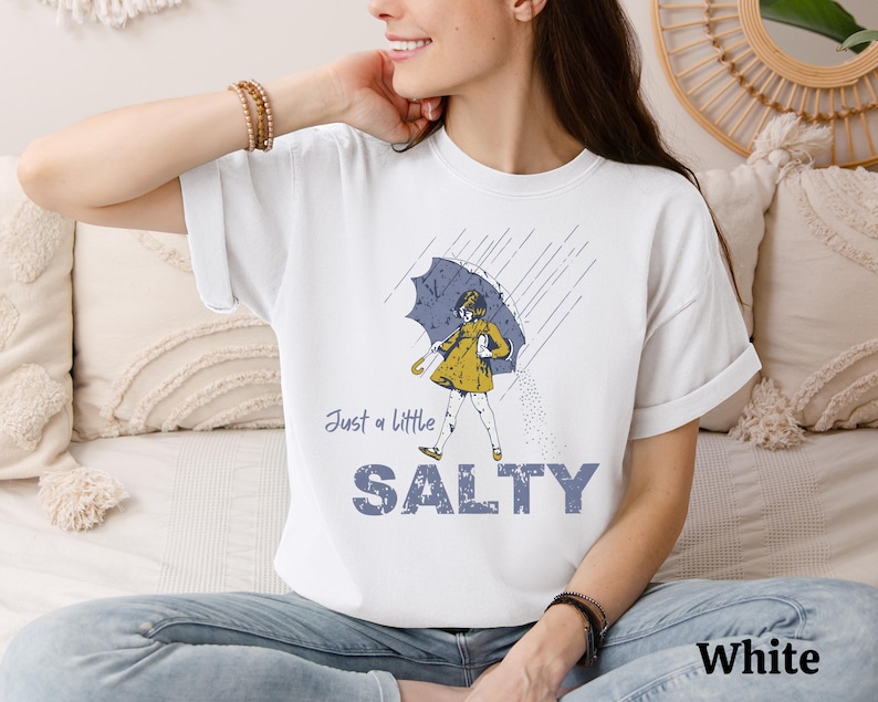 May include: White t-shirt with a graphic of a person under an umbrella in the rain. The text "Just a little SALTY" is below the graphic. The word "White" is printed at the bottom right.