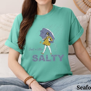 May include: Seafoam green t-shirt with the words "Just a little SALTY" in a purple font. The shirt features a graphic of a person in a yellow dress and umbrella in the rain. The shirt is a comfortable, casual style.