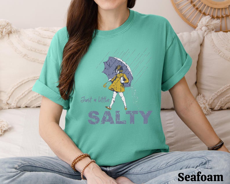 May include: Seafoam green t-shirt with the words "Just a little SALTY" in a purple font. The shirt features a graphic of a person in a yellow dress and umbrella in the rain. The shirt is a comfortable, casual style.