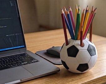 May include: A spherical pencil holder designed to resemble a black and white soccer ball, containing a collection of sharpened colored pencils in various shades including red, blue, green, yellow, and orange, positioned on a wooden desk.