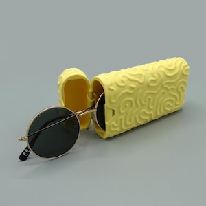 Könnte beinhalten: Runde Sonnenbrille mit goldenem Rahmen und dunklen Gläsern neben einem hellgelben, strukturierten Brillenetui. Das Etui ist geöffnet und gibt den Blick ins Innere frei. Das Etui und die Sonnenbrille befinden sich auf grauem Hintergrund.