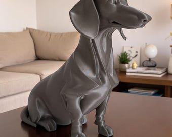May include: A gray dachshund dog statue, sitting upright with its head turned to the side. The sculpture has a smooth finish and is placed on a dark brown table. The dog's features are detailed, including its long ears and elongated snout.