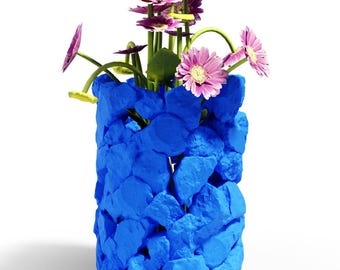 May include: A cylindrical vase constructed from numerous bright blue, rock-like textured pieces. The vase contains a bouquet of purple and pink gerbera daisies with green stems, some of which have small yellow circular accents on the stems.
