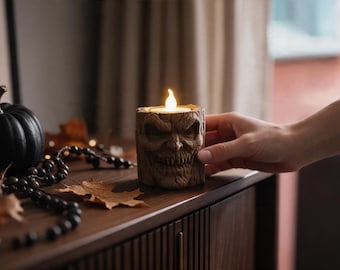 May include: A brown, cylindrical candle holder sculpted with a detailed, menacing face featuring deep-set eyes and a wide, toothy grin. The holder contains a small, glowing LED candle flame that emits a warm, soft light from the top center.