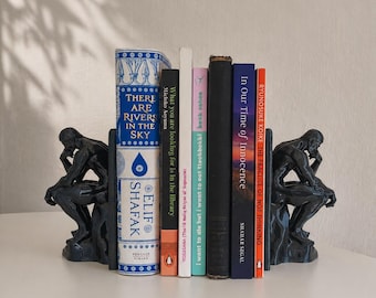 May include: Two black bookends shaped like The Thinker sculpture holding a row of six books. The books include titles such as There Are Rivers in the Sky by Elif Shafak, What You Are Looking For Is in the Library, and In Our Time of Innocence.