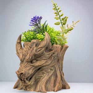 Puede incluir: Una maceta marrón con forma de dragón, llena de vegetación artificial y flores moradas y verdes. La maceta tiene un diseño detallado y texturizado que se asemeja a la madera. La cabeza del dragón se destaca, con cuernos y una cara detallada.